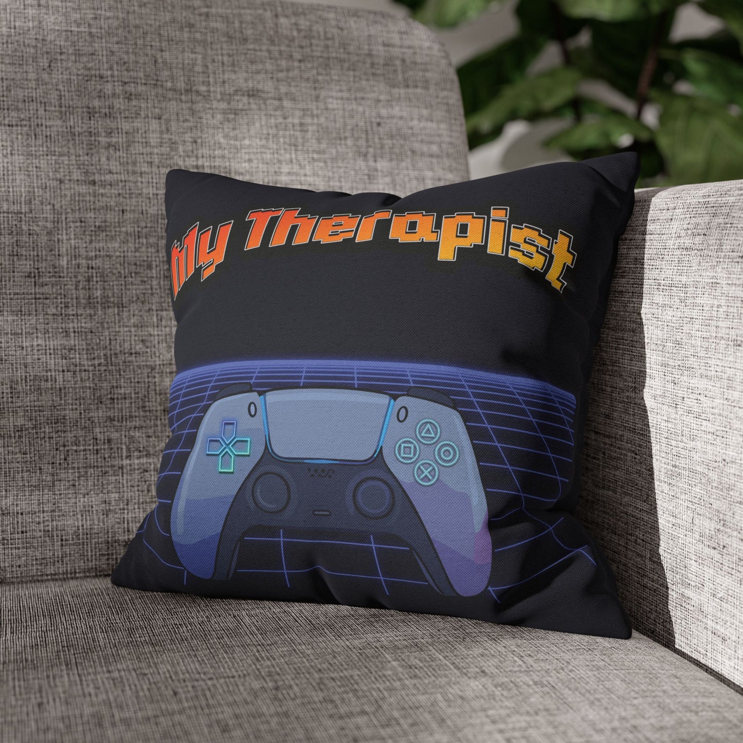 A square throw pillow resting on a gray fabric sofa. The pillow is black with the text 'My Therapist' in orange and yellow, above a stylized video game controller graphic in shades of blue, purple, and gray.
